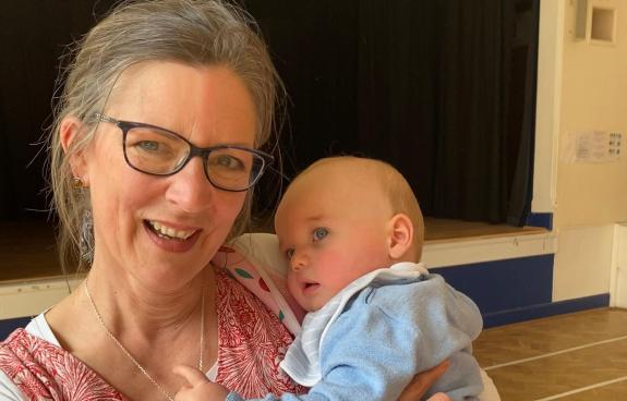A woman in her fifties is smiling and holding a baby boy who looks very happy and content. The woman is looking at the camera and the baby is reaching for her necklace.